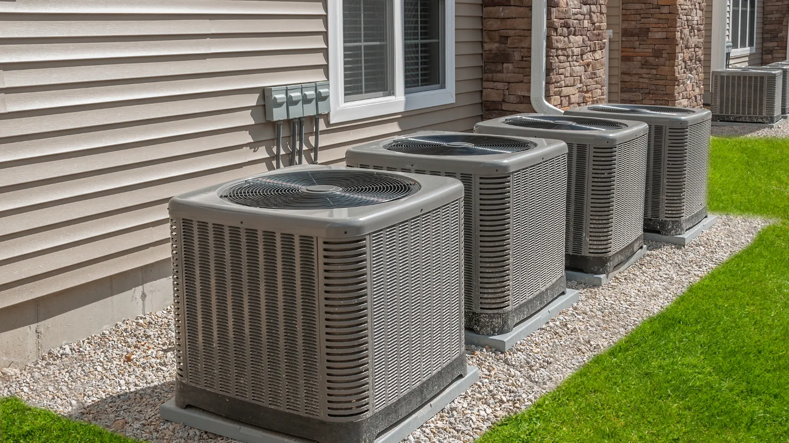 Four residential condensing units lined up on a gravel bed along the side of a house, with disconnect boxes on the wall above them.