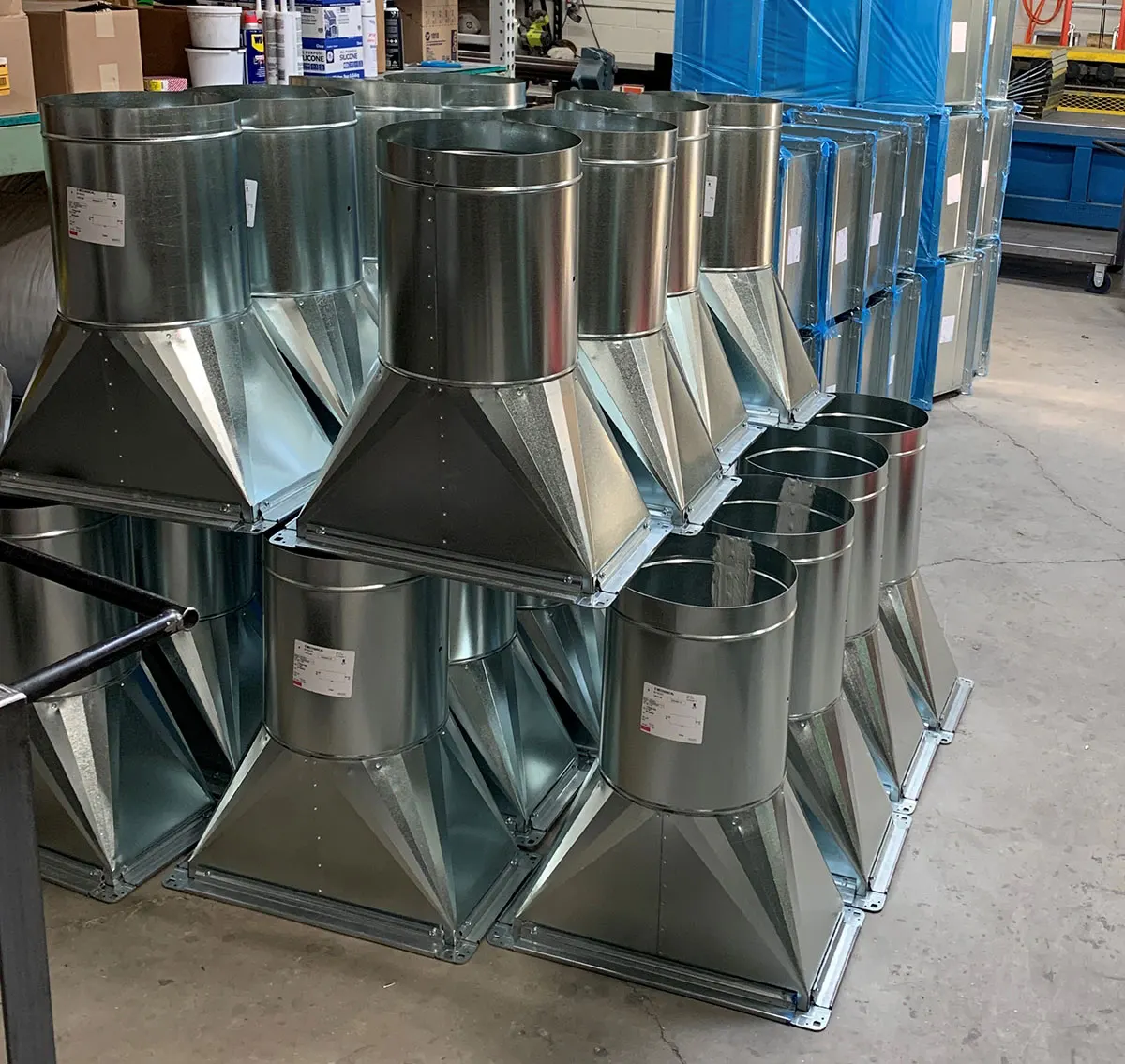 Rows of galvanised sheet metal boots and round takeoffs stacked on a shop floor, with blue-filmed panels racked behind them.
