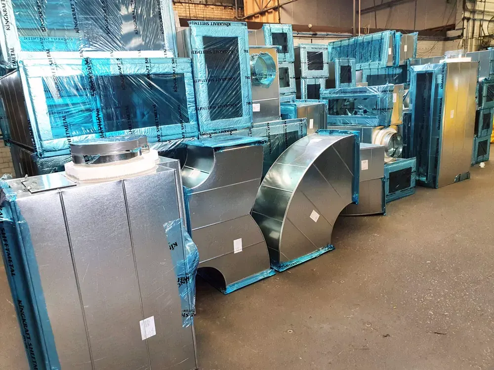 Fabricated galvanised sheet metal plenums and transitions stacked in a shop, most still wrapped in blue protective film.