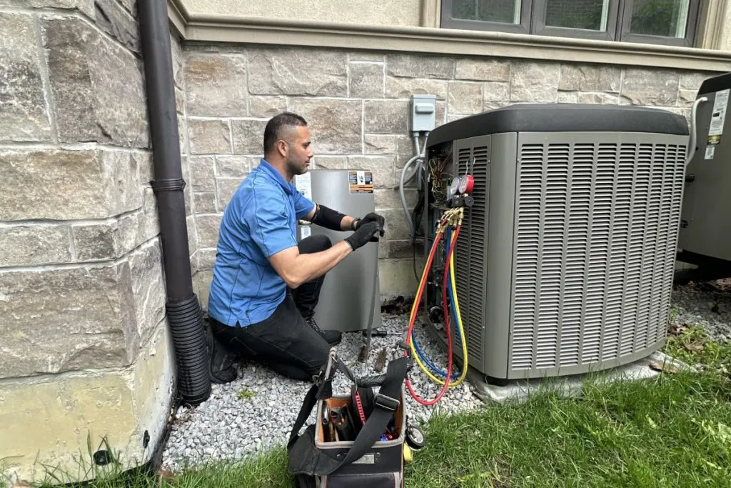 A technician in a blue polo kneeling at a residential condenser with the service panel off and a gauge manifold connected, tool bag open beside him.