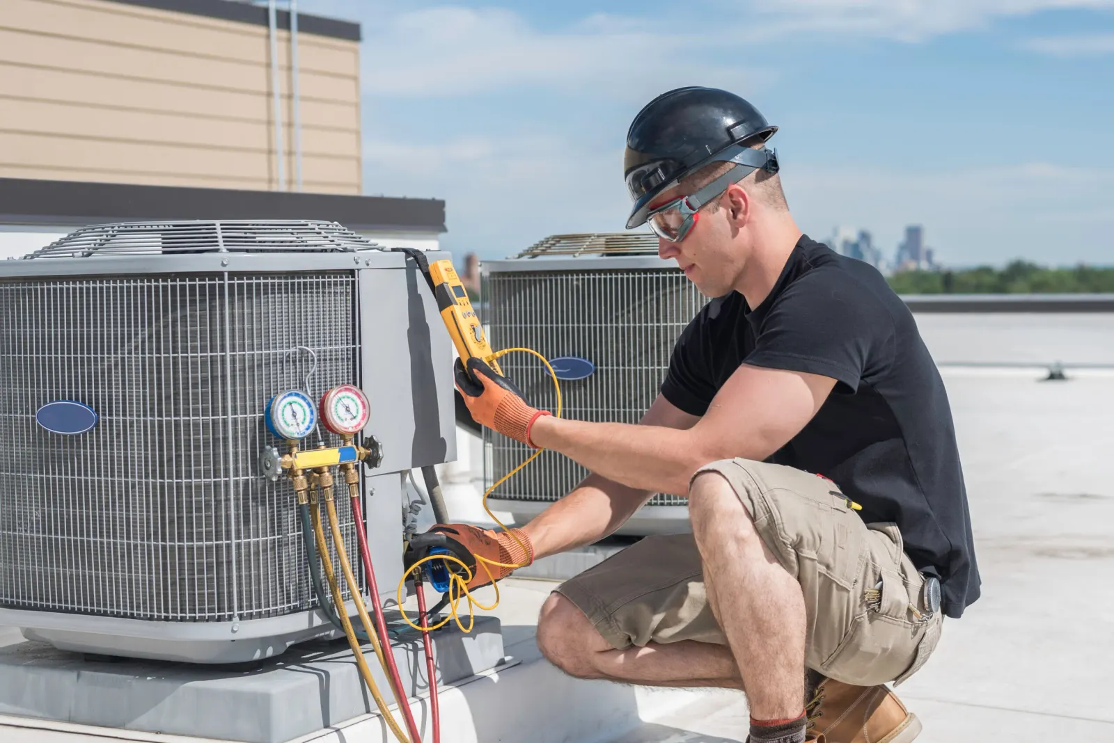 A technician in safety glasses and a hard hat crouched beside rooftop condensing units, taking a reading with a yellow multimeter.
